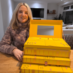 Smiling woman sitting at a table with the Selfridges 2025 Beauty Advent Calendar displayed in front of her, showing multiple stacked yellow gift boxes.