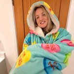 Woman wearing a colourful oversized Oodie hoodie, smiling with arms folded across her chest, standing indoors against a wooden door.